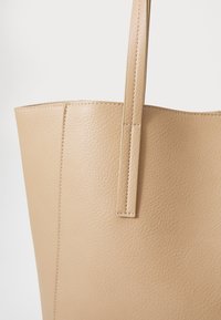 Sac fourre-tout en simili cuir beige avec une texture lisse, une forme structurée et une seule longue poignée, présentant des coutures visibles le long des bords.