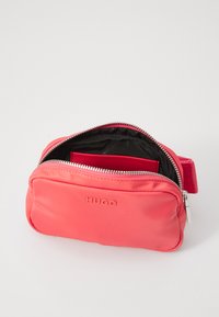 Sacoche en nylon rouge à porter en bandoulière, avec une fermeture zippée, arborant un logo sur le devant et un intérieur noir contrastant. Design compact et épuré.