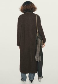 Osoba s kudrnatými vlasy, oblečená v dlouhém hnědém kabátu, modrých džínech a na podpatcích, která nese ramenní tašku ze které visí šály.