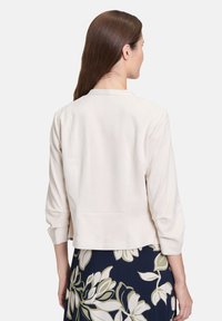 Jasno beżowa kurtka typu cropped z rękawami o długości 3/4, gładkiego materiału, prostym kołnierzem i bez widocznych zapięć, połączona z wzorzystą spódnicą.