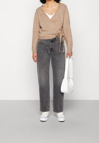 Maglione avvolto beige con dettagli a cravatta, indossato sopra una maglietta bianca, abbinato a jeans grigi a gamba dritta e sneaker bianche, con una piccola borsa bianca.