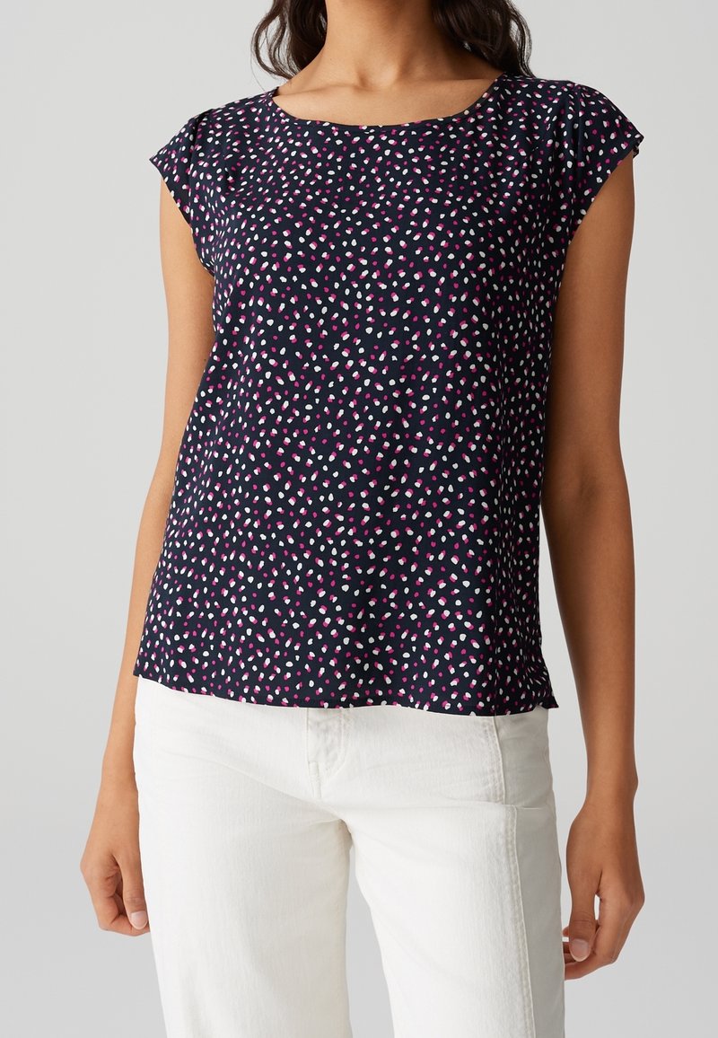 Femme portant un chemisier bleu marine à manches courtes avec un motif de points blancs et roses, associé à un pantalon blanc sur un fond uni.