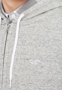 Primo piano di una felpa grigia con zip e cordini bianchi indossata sopra una camicia con colletto a scacchi.
