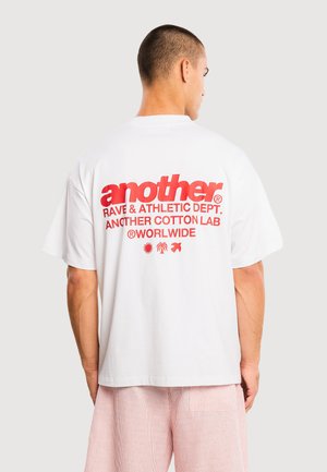 Mężczyzna w białym, oversize’owym T-shircie z czerwonym napisem „another” i tropikalnymi ikonami na plecach, w różowych szortach w paski, zwrócony tyłem.