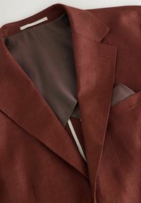 Blazer marron pour homme avec col à revers cranté et poche poitrine, montrant une doublure intérieure lisse et une texture de tissu structurée.