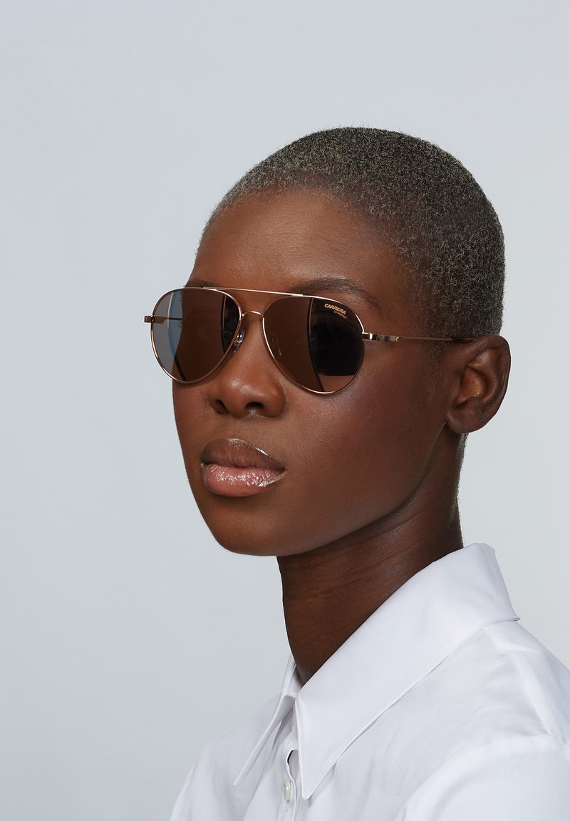 Sonnenbrille mit braun getönten Gläsern, Metallrahmen und runder Form. Getragen von einem Model mit kurzen grauen Haaren und einem weißen Hemd mit Kragen.