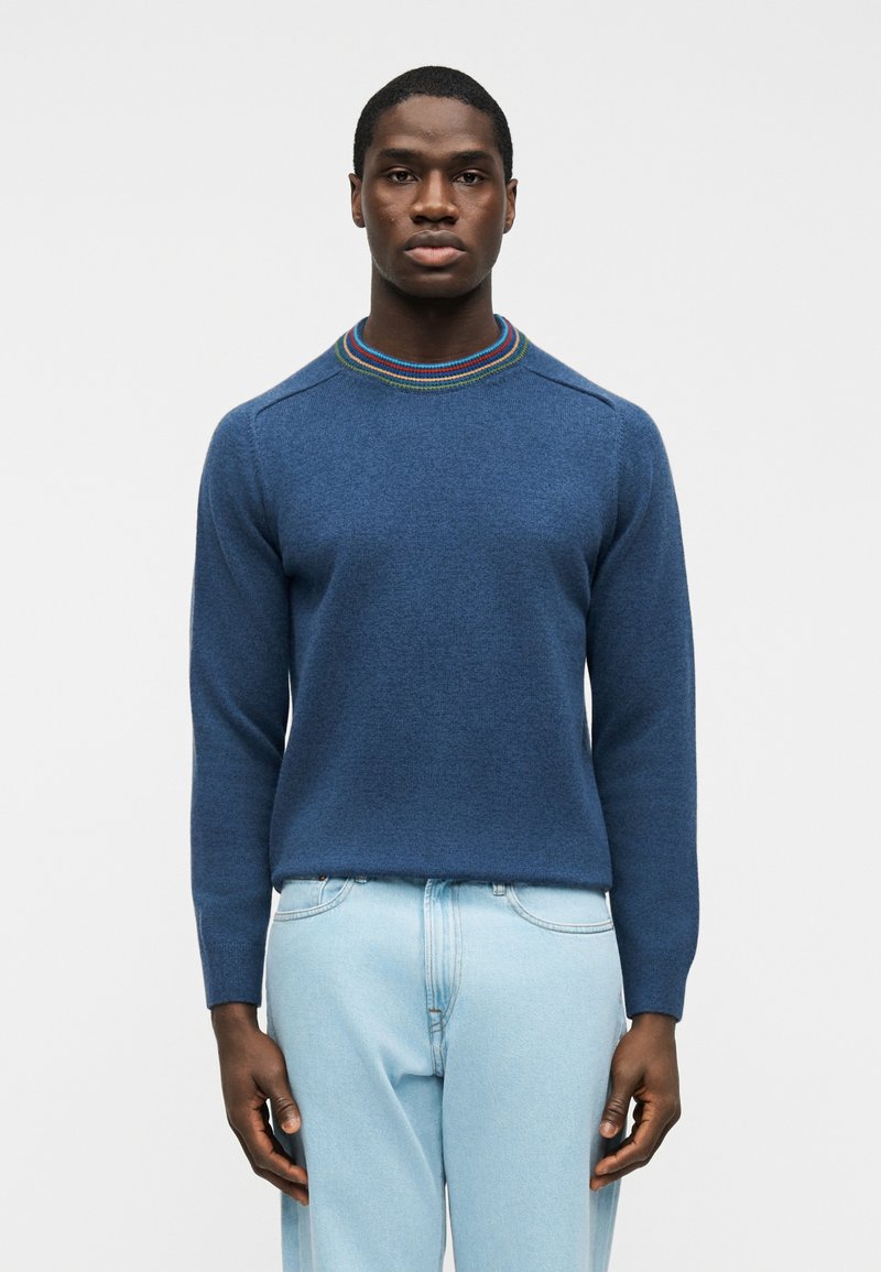 Homme portant un pull bleu avec un col rayé multicolore et un jean bleu clair, debout devant un fond blanc uni.