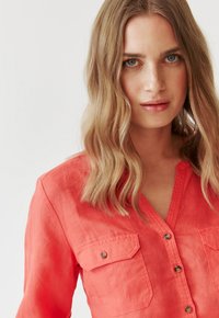 Camicia corallo con bottoni caratterizzata da due tasche sul petto, scollatura a V e un tessuto leggero e testurizzato. I bottoni e le cuciture sono visibili.