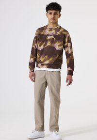 Braun-gelb gemusterter Tie-Dye-Pullover mit Rundhalsausschnitt, getragen über einem weißen Hemd, kombiniert mit hellbeigen Hosen und weißen Sneakers.