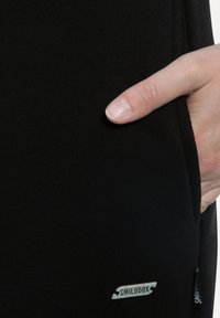 Schwarzer gerippter Stoff mit einer Seitentasche. Mit einem kleinen weißen Etikett mit der Aufschrift "SMILODOX" am Saum. Glatte Textur, enganliegendes Design.