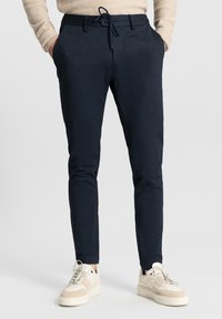 Marineblauwe tapered broek met een elastische tailleband, kleine zakken en een gestructureerde stof; gecombineerd met beige sneakers.