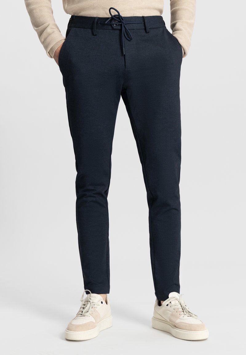 Marineblauwe tapered broek met een elastische tailleband, kleine zakken en een gestructureerde stof; gecombineerd met beige sneakers.