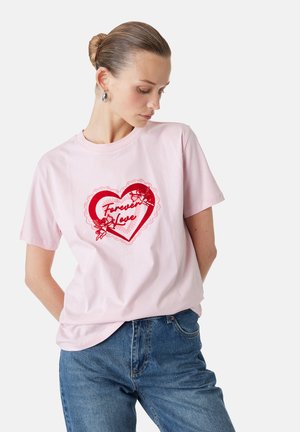 Kobieta z włosami upiętymi w kok nosi jasnoróżowy t-shirt z czerwonym sercem i napisem „Forever Love”, połączony z niebieskimi dżinsami, pozując na białym tle.