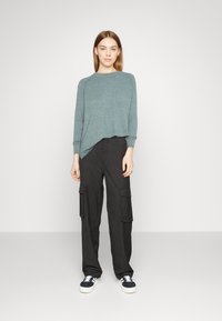 Maglione oversize a maniche lunghe di un leggero color teal abbinato a pantaloni cargo neri a righe con tasche laterali e sneakers con accenti bianchi.