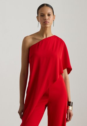 Rød jumpsuit med én skulder og drapert design. Glatt stoff og asymmetrisk kant. Tilbehør med en bred mansjettarmbånd på det ene håndleddet.