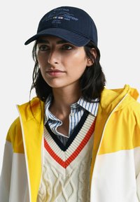 Cappello da baseball blu navy con testo ricamato, abbinato a una giacca con cappuccio gialla e un maglione bianco a trecce con scollatura a V multicolore.