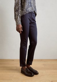Marineblå slim-fit bukser med tekstur, knaplukking og slits ved anklene, sammen med mørke loafers på et tregulv.