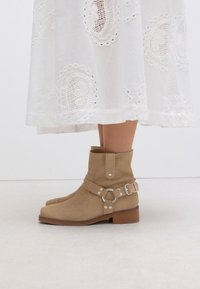 Helle Wildleder-Stiefeletten mit klobigem Absatz, ausgestattet mit einem dekorativen Riemen mit Schnalle und Metallring-Akzent. Glatte Textur und abgerundetes Zehendesign.