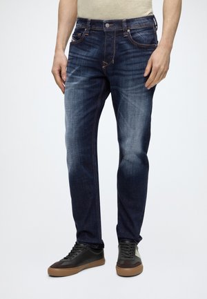 Homme portant un jean bleu foncé délavé et des baskets noires à semelles marron sur un fond clair uni.