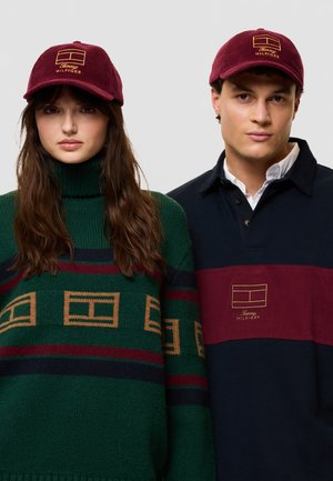 Dos modelos llevan gorras burdeos de Tommy Hilfiger; la mujer viste un jersey de rayas verdes y el hombre, un polo azul marino y burdeos.