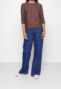 Donna che indossa un maglione a righe marroni e nere a maniche lunghe, jeans blu a gamba larga e sneakers bianche, posando su uno sfondo bianco uniforme.