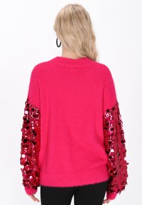 Bolondos, élénk rózsaszín pulóver hosszú ujjal, piros flitterekkel díszítve, laza fazonban és bordás kötött mandzsettával. Puha tapintású az egész anyag.