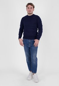 Maglione blu navy con scollo a giro, abbinato a jeans blu chiaro. Sneakers bianche completano l'outfit. Lo sfondo è semplice e di un colore chiaro.