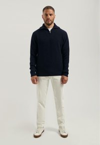 Marineblauw gebreid trui met een kwart-rits kraag, voorzien van ribbeltextuur. Combinatie met lichtgekleurde, rechte broek en witte sneakers.