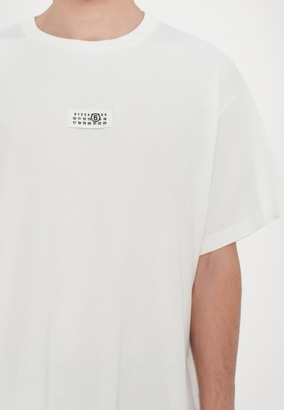 T-shirt en coton blanc à manches courtes, avec une étiquette rectangulaire noire comportant des chiffres et le numéro "6" affiché en évidence.