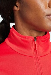 Giacca rossa con zip e colletto alto. Tessuto liscio con una vestibilità aderente, dotata di una robusta zip nera e un tirazip rosso sul collo.