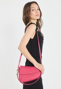 Rosa texturerad axelväska med klaffstängning och lång rem, bärs av en person i en svart outfit, med en slät yta.