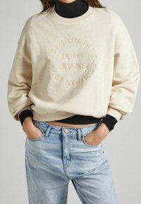Beige sweatshirt met reliëf logo, ronde halslijn en geribbelde zwarte manchetten, gecombineerd met lichtblauwe denim jeans.