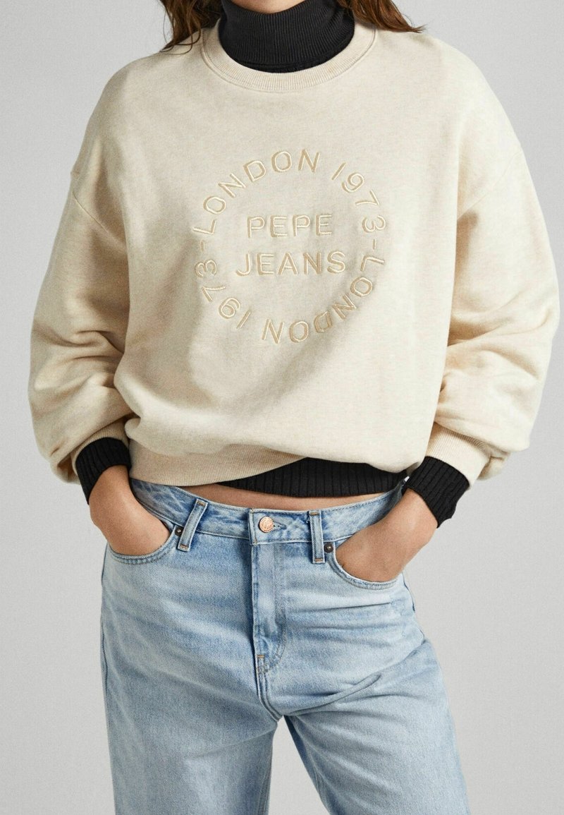 Beige sweatshirt met reliëf logo, ronde halslijn en geribbelde zwarte manchetten, gecombineerd met lichtblauwe denim jeans.