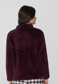 Chaqueta de forro polar burdeos con una superficie suave y texturizada, un cuello alto y mangas largas. Usada sobre un pantalón de patrón a cuadros.