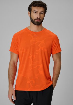 Mężczyzna z brodą w jasnoróżowej pomarańczowej koszulce z krótkim rękawem i subtelnym abstrakcyjnym wzorem, stojący na jednolitym szarym tle.