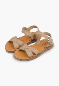 Beige-nahkasaappaat ristikkäisillä hihnoilla, säädettävällä soljella, littealla ruskealla pohjalla ja vaaleilla ompeluyksityiskohdilla.