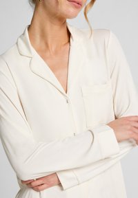 Crème-kleurig pyjamajasje met lange mouwen, een kraag en een borstzak. Gemaakt van zachte stof met knoopsluitingen.
