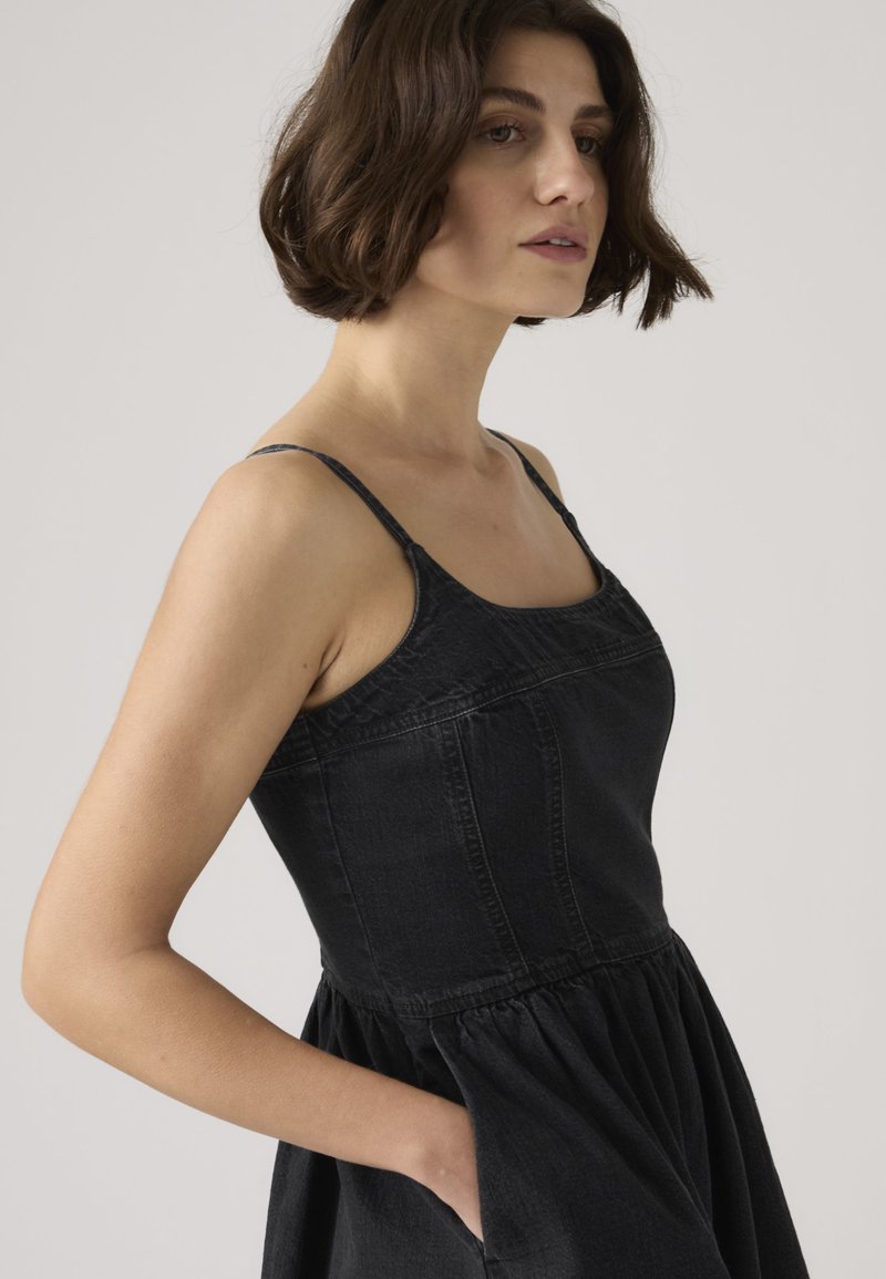 Vrouw met kort bruin haar die een mouwloze zwarte spijkerjurk draagt met een nauwsluitend lijfje en een geplooide rok, hand in zak, kijkend naar links.