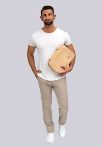 Bolsa de portátil color marrón claro con un acabado suave, diseño espacioso y asa superior, sostenida por una persona con una camisa blanca y pantalones gris claro frente a un fondo neutro.