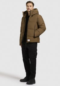 Brun pufferjakke med hætte, frontlynlås og to sidelommer med lynlås. Kombineret med sorte cargo-bukser og sorte snøre støvler.