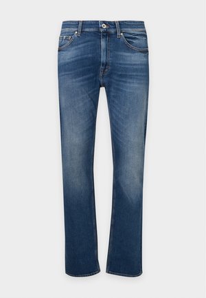 Mand stående med hænder delvist i lommerne iført en sort t-shirt, blå jeans og beige sneakers mod en ensfarvet lys baggrund.