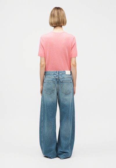 Femme aux cheveux blonds courts portant un t-shirt rose à manches courtes et un jean large bleu surdimensionné, vue de dos sur un fond blanc.