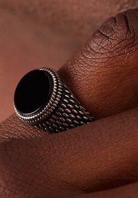 Metallring mit einem glatten schwarzen Stein in der Mitte und einem strukturierten Silberband mit einem Netzmuster. Auf einem dunklen Hautton positioniert.