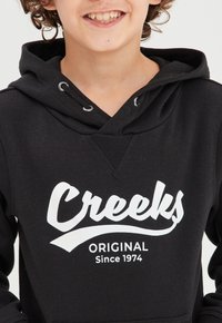 Felpa nera con scollo a V, caratterizzata da un logo bianco "Creeks" e la scritta "Original Since 1974". Il materiale sembra morbido e testurizzato.