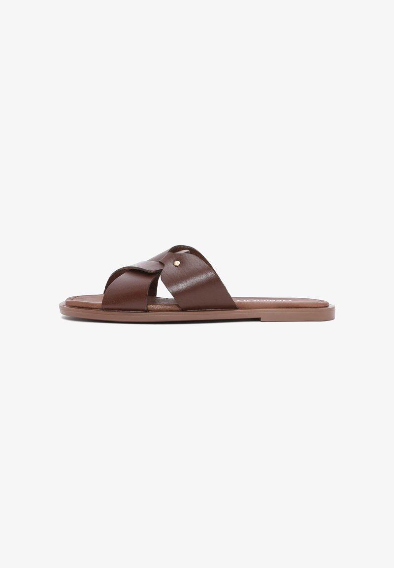 Sandale plate marron avec de larges sangles en cuir croisées et un petit clou en métal sur un fond blanc uni.