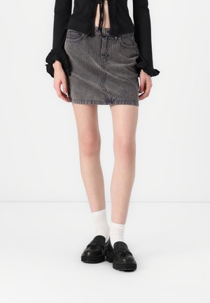 Kobieta ubrana w czarny krótki top, wyblakłą czarną mini spódnicę z denimu, białe skarpetki i czarne mokasyny z frędzlami, stojąca na tle jednolitego białego tła.