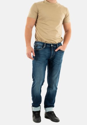 Camiseta de manga corta beige combinada con jeans azul oscuro con dobladillo vuelto. El modelo lleva zapatillas negras y está de pie con una mano en el bolsillo.