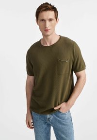 Oliwkowa, krótko-sleeve koszulka z okrągłym dekoltem i kieszonką na piersi. Materiał ma strukturalne wykończenie. Noszona z jasnoniebieskimi dżinsami.