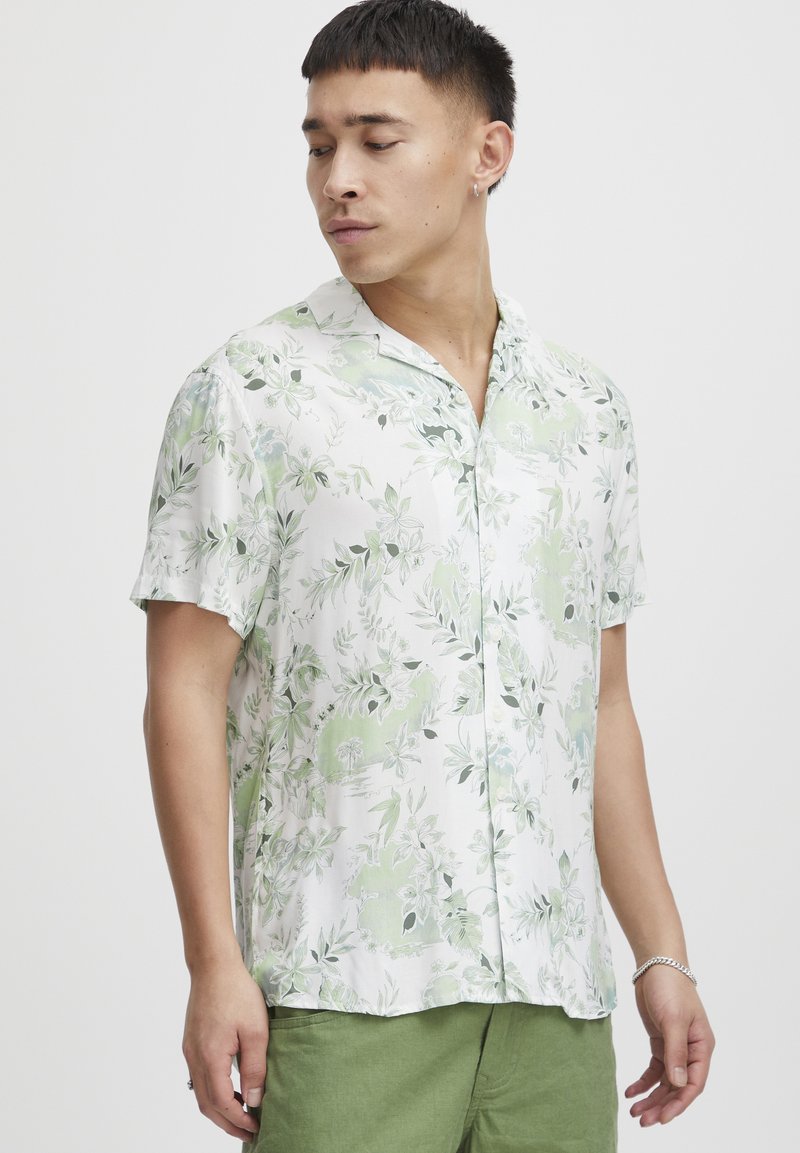 Camisa de flores blanca de manga corta con hojas verdes, fabricada en tejido ligero, con cuello relajado y cierre de botones, que presenta un patrón sutil y una textura suave.