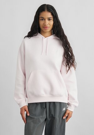 Young woman with long curly hair wearing a light pink hoodie and gray New Balance pants, standing against a plain light background.